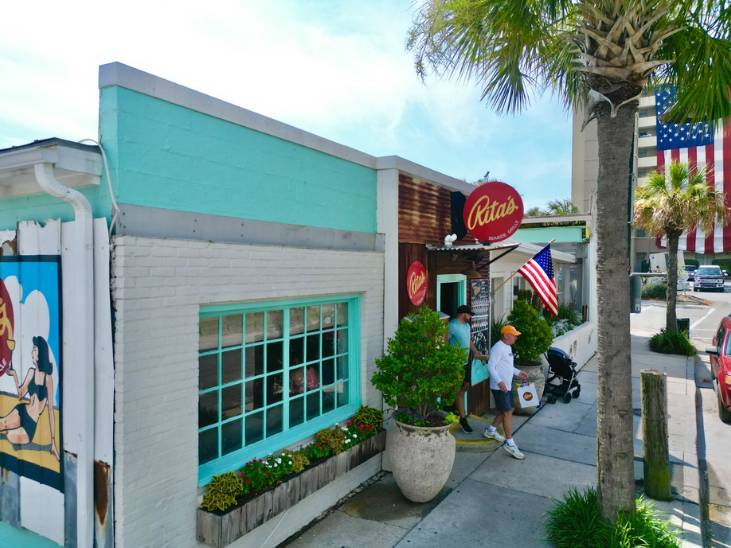 Exterior of Rita’s Seaside Grille with a turquoise and white facade, palm trees, and guests entering from the sidewalk.