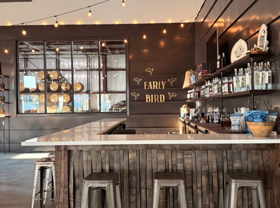 Modern distillery tasting room with a wooden bar, metal stools, shelves of spirits, string lights, and barrels visible behind glass.