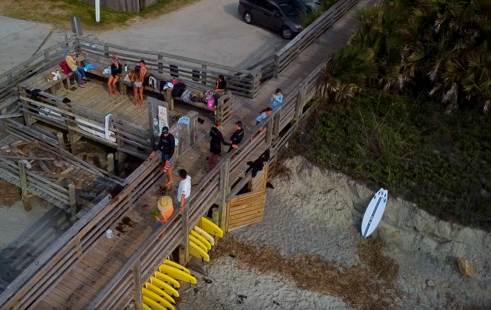 Aerial view of surfers gathering on a wooden beach access walkway, with yellow surfboards lined up beside the dunes.