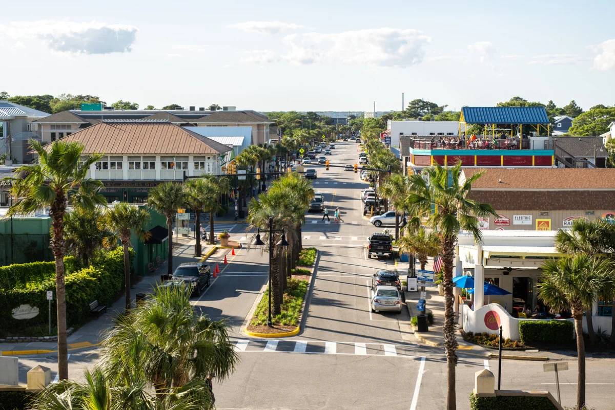 Palm-lined street in a coastal town with shops, restaurants, parked cars, and pedestrians under a bright sky.