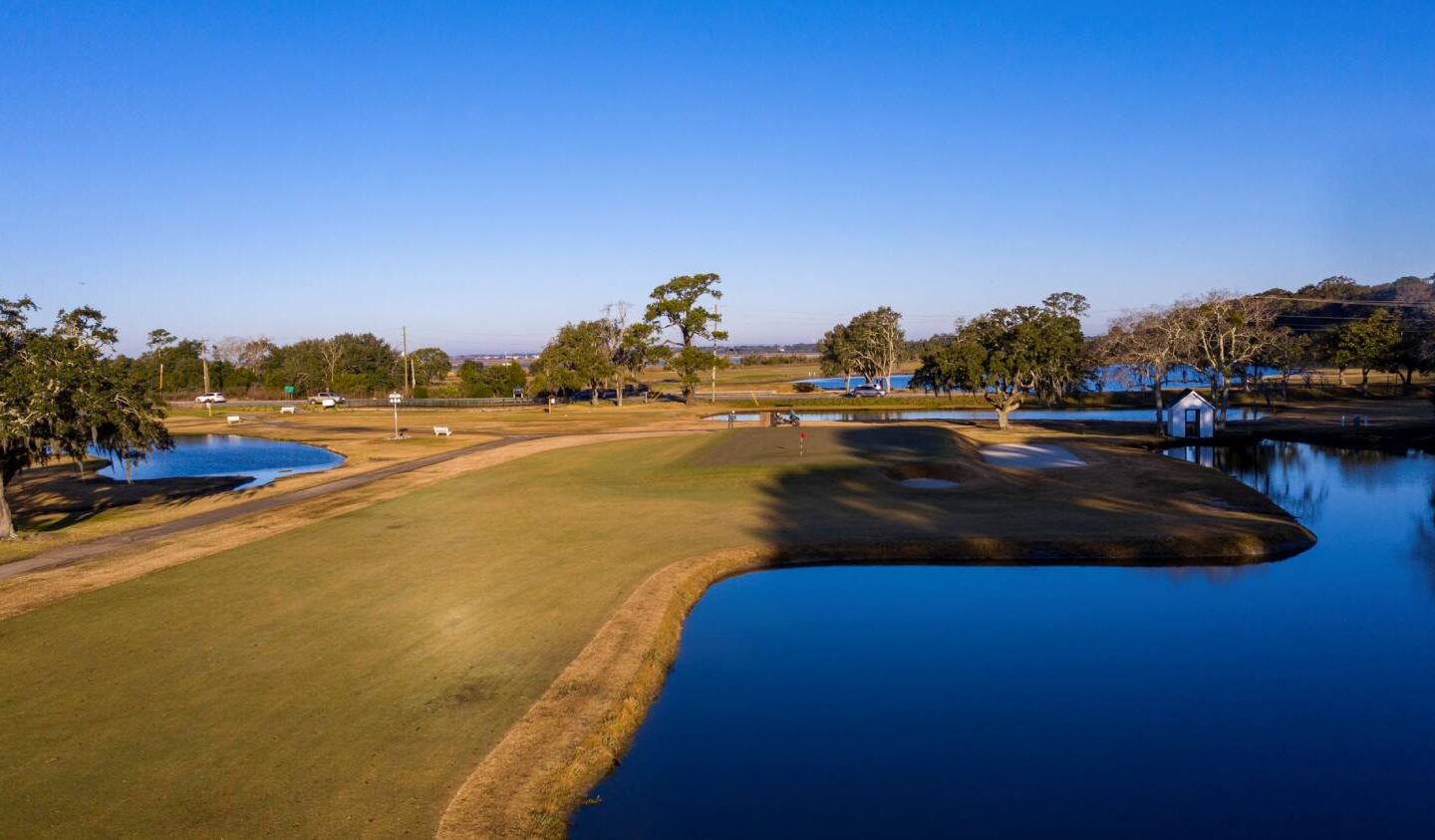 Aerial view of a golf course fairway surrounded by ponds, trees, and sand bunkers beneath a clear blue sky.