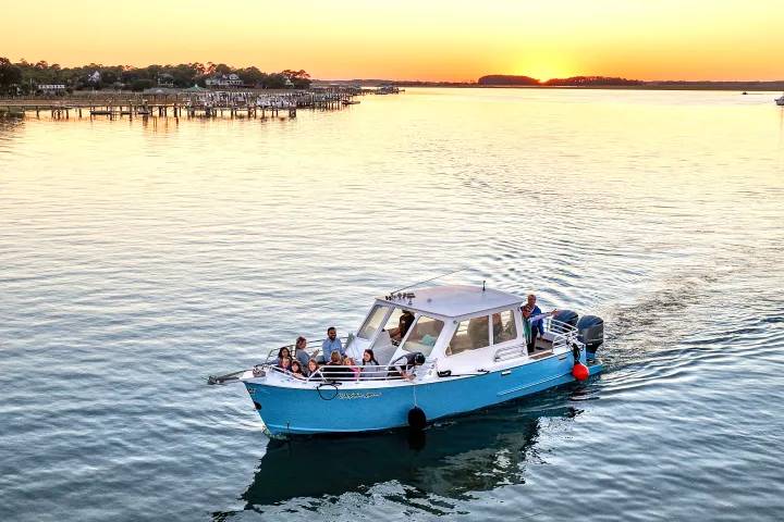 Boat tour with passengers cruising across calm water at sunset, with docks and waterfront homes in the distance.