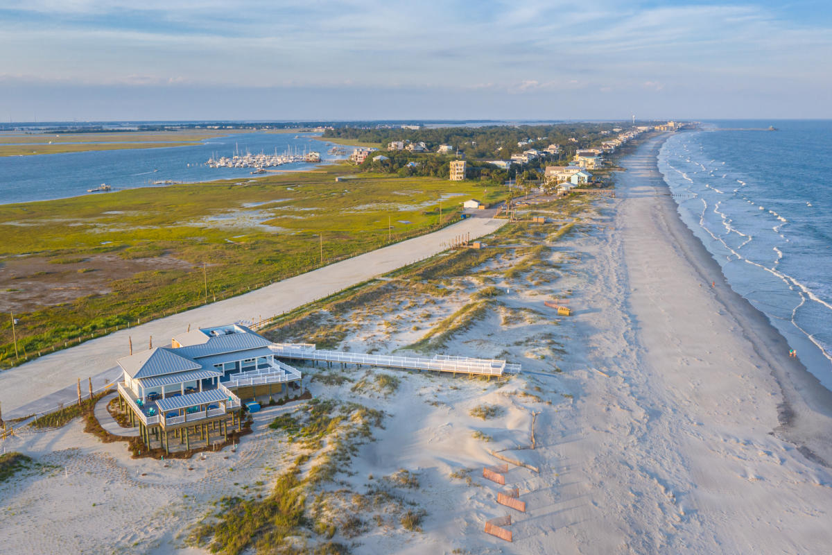 Aerial view of Folly Beach County Park with a beachfront pavilion, sandy shoreline, ocean waves, marshland, and a marina.