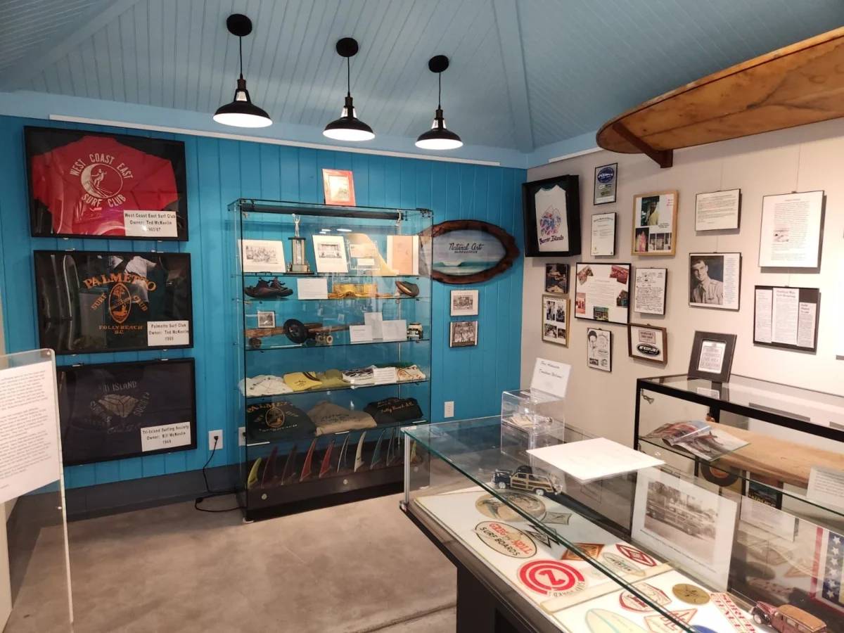 Interior of the Folly Beach History Museum with surf memorabilia, vintage photographs, framed documents, and artefacts displayed in glass cases.