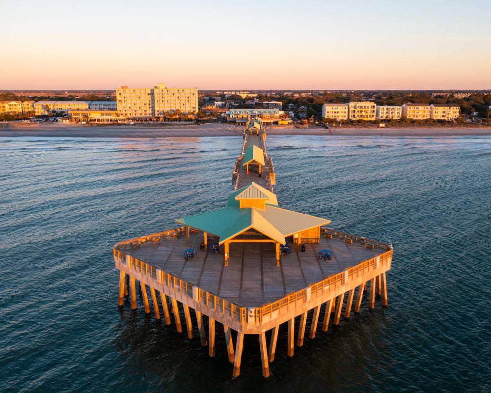 Aerial view of the Folly Beach Fishing Pier extending into the ocean, with beachfront hotels and buildings glowing in the evening light.