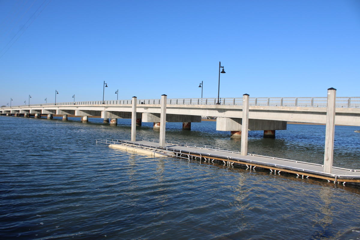 Boat landing with a floating dock beside a concrete bridge over calm blue water under a clear sky.