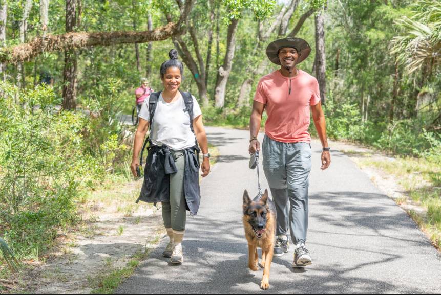 Two hikers walking a German Shepherd on a paved forest trail surrounded by lush greenery.