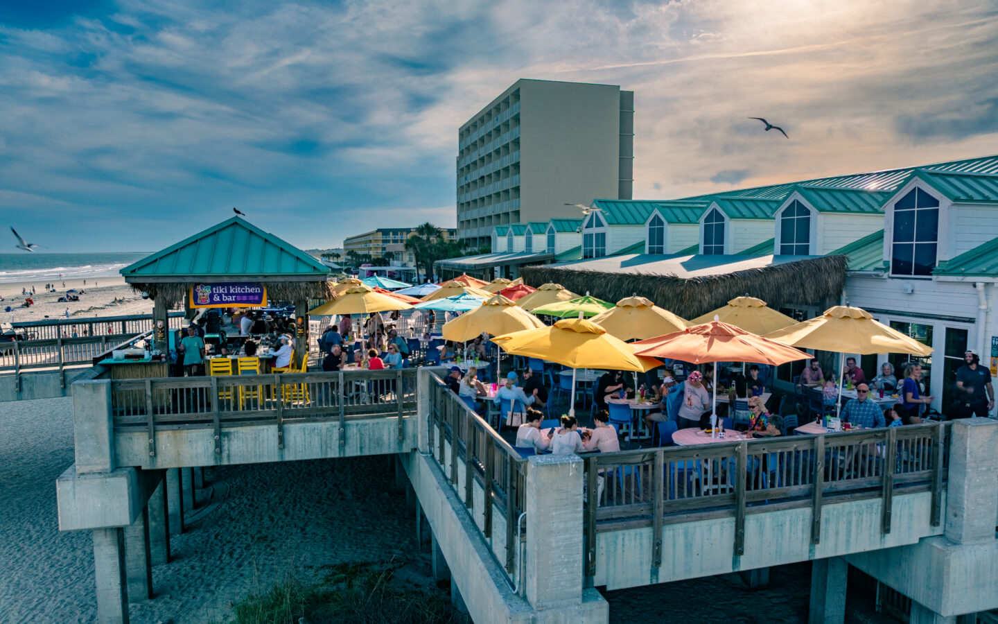 Beachfront restaurant patio filled with diners beneath colourful umbrellas, overlooking the ocean beside a hotel.