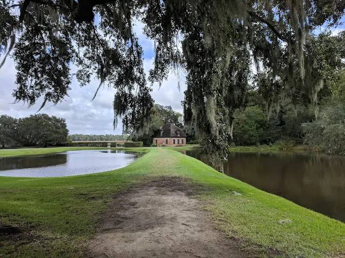 Scenic path beside a pond leading to a historic brick building, framed by large oak trees draped in Spanish moss.