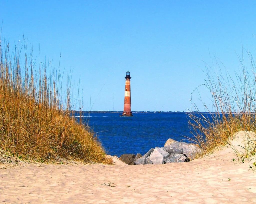 Morris Island Lighthouse viewed across the water from a sandy beach framed by dune grass and rocks.