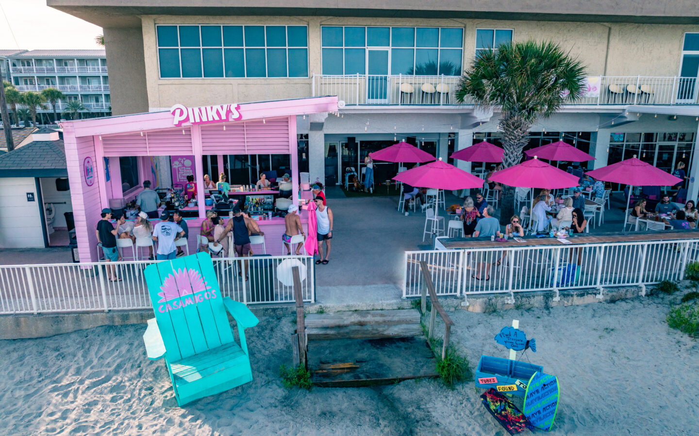 Beachfront bar and patio with bright pink umbrellas, outdoor seating, and guests gathered beside the sand.