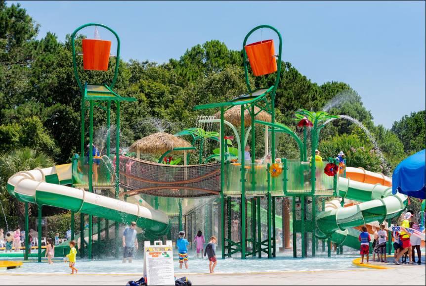 Children playing on a colourful outdoor water playground with slides, spraying fountains, and large tipping buckets.